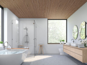 Un bagno moderno e luminoso con ampie piastrelle da bagno grigie, dotato di una vasca freestanding, una doccia in vetro, e una doppia vanità, il tutto sottolineato da un soffitto in legno a listelli e una vista sulla natura attraverso una grande finestra.