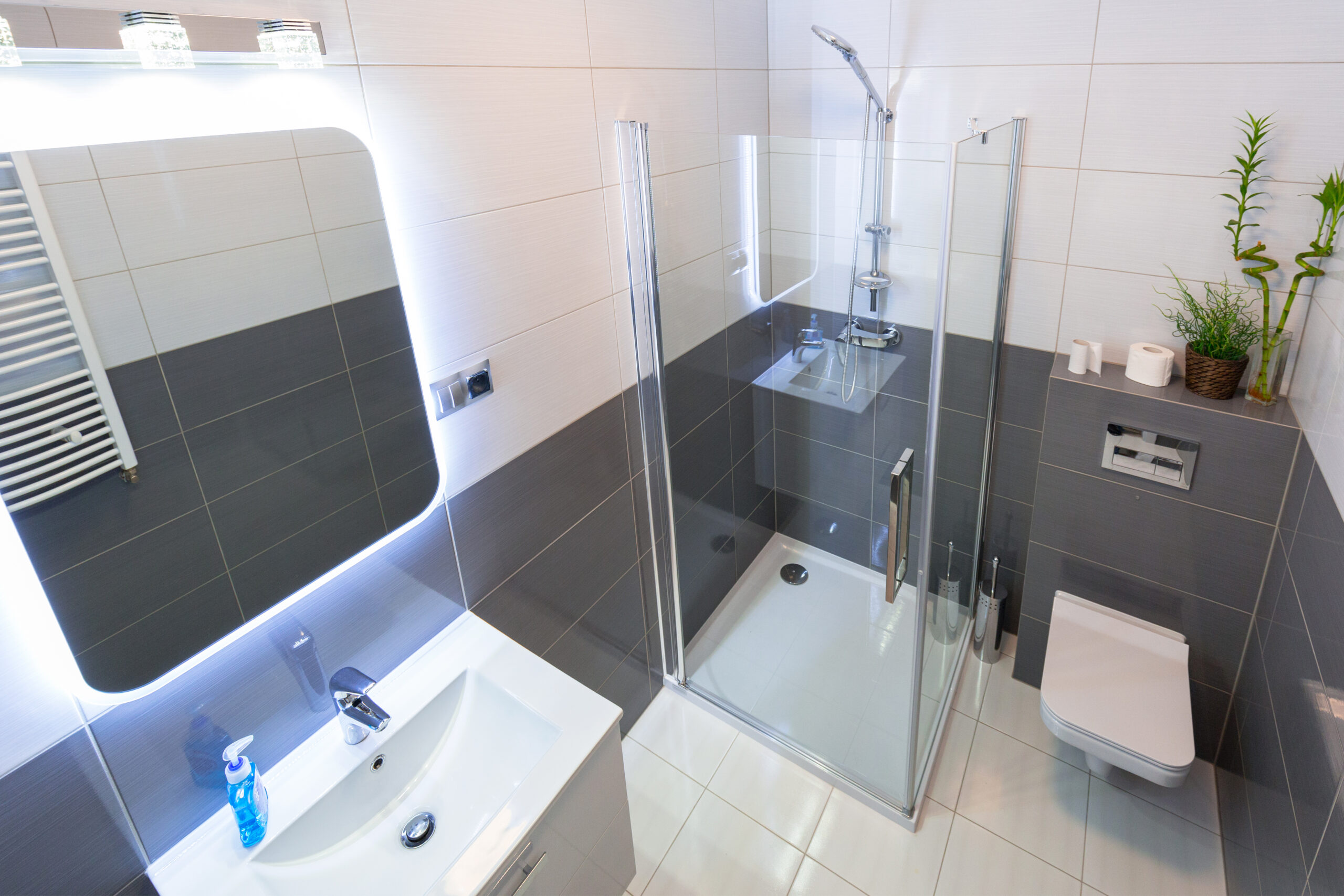 Un bagno moderno e ristrutturato senza opere murarie, dotato di piastrelle bicolori, una cabina doccia in vetro, sanitari sospesi e un specchio illuminato, il tutto progettato per massimizzare lo spazio e il comfort.
